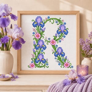 May include: A square wooden frame containing a cross-stitch pattern of the letter R, intricately decorated with purple iris flowers, green vines, and small pink blossoms on a white grid fabric background, displayed as a piece of wall art.