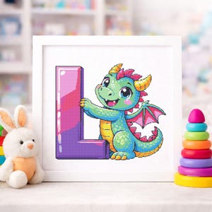 Könnte beinhalten: Ein gerahmtes Kreuzstichbild mit einem freundlichen Zeichentrickdrachen, der den Buchstaben "L" hält. Der Drache ist grün, blau und gelb mit rosa Akzenten. Der Buchstabe ist lila und rosa. Ein Plüschhase und ein Stapel bunter Ringe sind im Vordergrund.