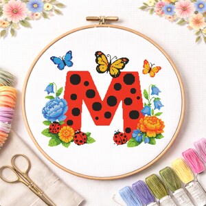 Könnte beinhalten: Kreuzstich-Stickerei in einem Holzrahmen mit einem roten Buchstaben "M" mit schwarzen Punkten, umgeben von Schmetterlingen, Marienkäfern und floralen Elementen in Blau, Gelb und Orange. Das Design ist vor einem weißen Hintergrund.
