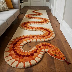 Puede incluir: Una alfombra beige y naranja con forma de serpiente y un patrón de escamas detallado. La alfombra está colocada sobre un suelo de madera, con la cabeza y la cola de la serpiente extendiéndose más allá del cuerpo principal de la alfombra. El diseño añade un elemento decorativo único.