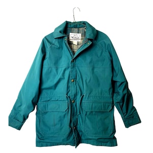 May include: A teal blue jacket with a plaid lining and a button closure. The jacket has two large pockets on the front and a drawstring at the waist. The label on the jacket reads "The Woolrich Company".