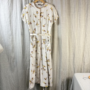 Könnte beinhalten: Ein weißes Vintage-Kleid mit einem Blumenmuster in Gelb und Braun. Das Kleid hat kurze Ärmel, eine Knopfleiste vorne, einen passenden Gürtel und Taschen. Das Kleid hängt an einem Holzkleiderbügel.
