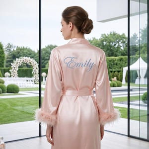 May include: A light pink satin robe with feather trim on the sleeves and the name "Emily" embroidered on the back. The robe is tied at the waist with a matching belt. The background shows a wedding ceremony setup with a white floral arch.