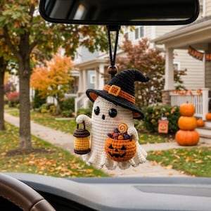 May include: A crocheted ghost ornament hanging from a rearview mirror. The ghost wears a black witch hat with an orange band, holds a small brown lantern, and carries an orange jack-o'-lantern bucket filled with colorful candy pieces.