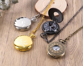 May include: Four pocket watches with chains are displayed on a wooden surface. One is silver with engraved initials, another is gold with a deer design and name, a third is black with an open lid, and the last is bronze with Roman numerals.