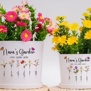 May include: Two white ceramic flower pots with a floral design and personalized text. The pot on the left says "Nana's Garden Love Grows Here" and the pot on the right says "Nana's Garden Make Life More Grand".