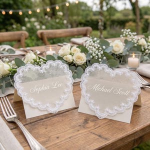 Op de afbeelding: Twee hartvormige naambordjes met witte kanten rand, elk met een naam, liggen op een houten tafel. De tafel is gedekt voor een bruiloft, met bloemen, kaarsen en bestek. De namen op de kaarten zijn "Sophia Lee" en "Michael Scott."
