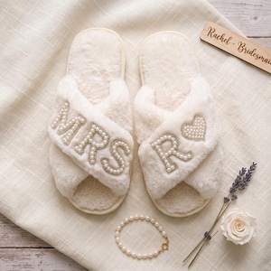 May include: A pair of white faux fur slippers with crisscross straps. The left slipper features the letters MRS in white pearls, and the right slipper features the letter R and a heart shape in white pearls. A wooden tag reads Rachel - Bridesmaid.