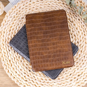 Könnte beinhalten: Zwei Tablet-Hüllen aus Leder, eine braune und eine dunkelgraue, mit Krokodilhautstruktur. Die braune Hülle trägt den Namen "Kate" in goldfarbenen Buchstaben. Die Hüllen liegen auf einer gewebten Matte.