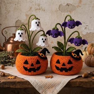 May include: Two crocheted orange pumpkin pots with black jack-o'-lantern faces and small black witch hats. One pot features white ghost-shaped flowers on green stems, while the other displays purple bat-shaped flowers on green stems.
