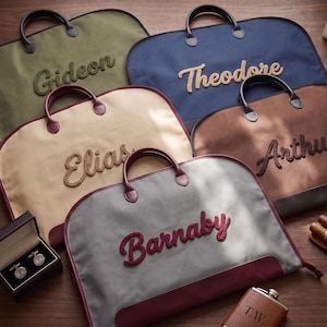 Puede incluir: Cinco fundas para ropa en verde oliva, azul marino, beige, marrón y gris con detalles en granate, cada una con un nombre bordado en letra cursiva texturizada: Gideon, Theodore, Elias, Arthur y Barnaby. Accesorios: caja de gemelos, petaca y puros.