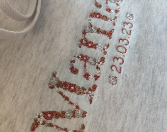 May include: A gray hoodie with the word "remarkable" embroidered in brown and white thread. The date "23.03.23" is embroidered below the word.