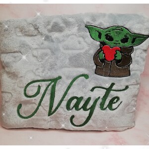 Peut inclure: Une couverture en peluche blanche avec un personnage brodé en vert tenant un cœur rouge et le nom "Nayte" brodé en vert.