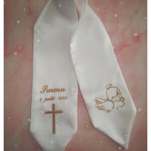 May include: White satin christening sash with gold embroidery. The sash features a cross, an angel, and the text "Swann 8 juillet 2023".