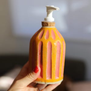 May include: A ceramic soap dispenser with a yellow and pink striped design. The dispenser has a white pump and a cork base. The bottle is approximately 15 cm tall.