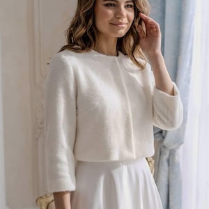 May include: A white cropped jacket with a soft, fuzzy texture, featuring three-quarter length sleeves and a round neckline, paired with a plain white skirt. The jacket has a clean, minimalist front closure and a structured yet comfortable fit.