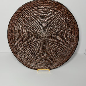 May include: A large, round decorative plate with a textured, concentric pattern in shades of brown and copper. The plate has a dark rim and is displayed on a gold-colored stand. The design gives the plate a rustic, handcrafted appearance.