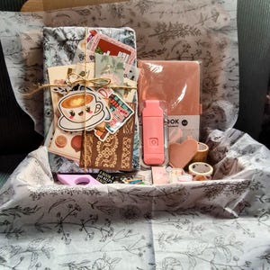 Puede incluir: Una caja de regalo llena de varios artículos, incluyendo una libreta rosa, una unidad USB rosa y una variedad de pegatinas y papel decorativo. La caja está forrada con papel de seda blanco con un patrón floral.