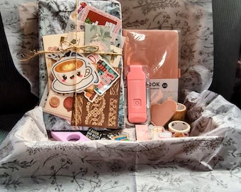 May include: A gift box containing stationery items such as a notebook, stickers, washi tape, and a pink water bottle. The items are wrapped in floral tissue paper and tied with twine. A coffee cup sticker is visible.