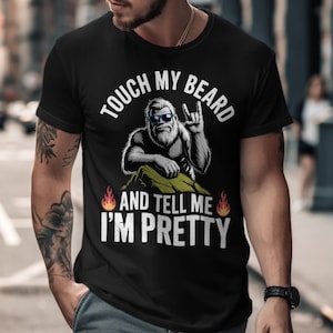 Puede incluir: Camiseta negra con un gráfico de un personaje parecido a Pie Grande con gafas de sol y haciendo el gesto de rock. El texto dice "TOUCH MY BEARD AND TELL ME I'M PRETTY" en blanco. Hay llamas a ambos lados del texto.
