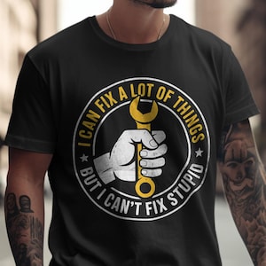Puede incluir: Camiseta negra con un diseño circular. El diseño muestra una mano agarrando una llave inglesa amarilla, rodeada por el texto "I CAN FIX A LOT OF THINGS BUT I CAN'T FIX STUPID." La camiseta tiene cuello redondo.