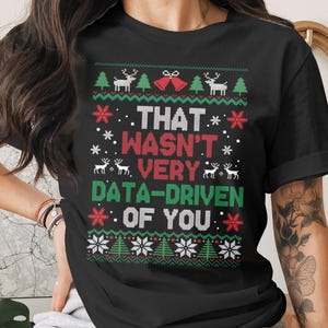 May include: Black t-shirt with a festive Christmas design. The shirt features a pattern of trees, snowflakes, and reindeer, with the text "THAT WASN'T VERY DATA-DRIVEN OF YOU" in white, red, and green.