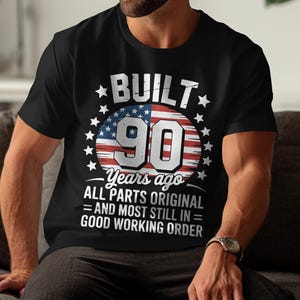 May include: Black t-shirt with a graphic that reads "BUILT 90 Years ago ALL PARTS ORIGINAL = AND MOST STILL IN = GOOD WORKING ORDER." The graphic features a red, white, and blue design with stars and the number 90.