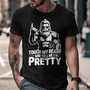 Puede incluir: Camiseta negra con un gráfico blanco de un Bigfoot musculoso con gafas de sol, señalando hacia arriba. El texto "TOUCH MY BEARD AND TELL ME I'M PRETTY" está impreso debajo en una fuente audaz y estilizada. La camiseta es de manga corta.
