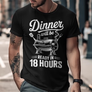 Pode incluir: T-shirt preto com um gráfico branco de uma churrasqueira e o texto "Dinner will be ready in 18 hours". O design inclui uma espátula e um garfo. A camisa tem gola redonda.