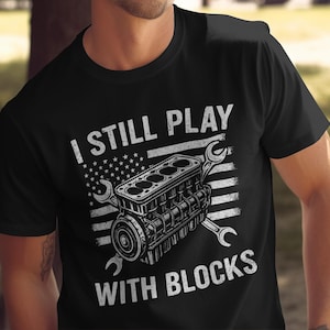 May include: Black t-shirt with a graphic of an engine block and wrenches, set against a distressed American flag design. The text reads "I STILL PLAY WITH BLOCKS" in white.
