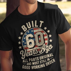 May include: Black t-shirt with a graphic design celebrating a 60th birthday. The design features the text "BUILT 60 Years Ago" in a circular emblem with the American flag and stars. Additional text reads "ALL PARTS ORIGINAL AND MOST STILL IN GOOD WORKING ORDER."