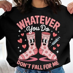 Puede incluir: Sudadera negra con un gráfico de dos calcetines de dibujos animados con caras, corazones y una jeringa. El texto dice "WHATEVER You Do DON'T FALL FOR ME" en rosa y blanco.