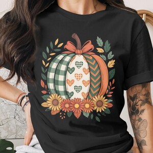 Peut inclure: T-shirt noir avec un motif de citrouille colorée. La citrouille présente des sections à carreaux verts, des chevrons orange et un centre blanc avec des motifs de cœurs. Ornée d'un nœud, de feuilles et de tournesols, parfaite pour l'automne.