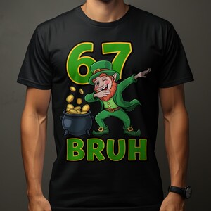 Peut inclure: T-shirt noir avec un motif de la Saint-Patrick. Le motif représente un dessin animé de lutin faisant un dab à côté d'un pot d'or, avec le chiffre 67 et le mot "BRUH" en vert et jaune.