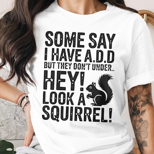 May include: White t-shirt with black text that reads "SOME SAY I HAVE A.D.D BUT THEY DON'T UNDER.. HEY! LOOK A SQUIRREL!" A black squirrel graphic is also featured. The shirt is a casual, humorous design.