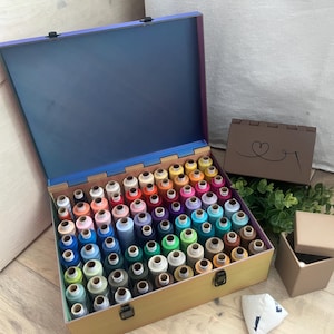 Puede incluir: Una caja de costura de madera abierta, llena de numerosos carretes de hilo colorido. La caja revela un espectro de colores vibrantes. También hay una caja marrón más pequeña con un diseño de corazón y una caja de cartón.