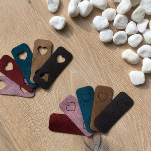 Puede incluir: Un conjunto de diez etiquetas de tela en forma de corazón de varios colores, incluyendo marrón, rosa, rojo, azul verdoso y negro. Las etiquetas están dispuestas en forma de abanico sobre una superficie de madera.