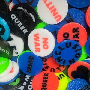 Könnte beinhalten: Nahaufnahme einer Sammlung farbenfroher, runder Token. Die Token zeigen Wörter wie „QUEER“, „UNITY“ und „NO WAR“ in fetter Schrift. Die Farben umfassen Rot, Blau, Grün, Schwarz und Weiß und erzeugen ein lebendiges Display.