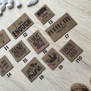 May include: A collection of brown square patches with various black text designs. Phrases include "I am enough," "Good Vibes Only," and "Perfectly Imperfect." Other patches feature a lightbulb, a unicorn, and a bee.