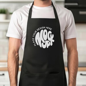 Op de afbeelding: Een persoon draagt een wit T-shirt en een zwarte schort met de tekst "Place your design here Mock Up" in wit.
