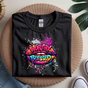 Op de afbeelding: Zwart t-shirt met een kleurrijk graffiti-ontwerp van lippen en de woorden "WORTHY BLESSED". Het ontwerp gebruikt heldere kleuren zoals roze, paars, blauw en geel, met verfspattereffecten. Het t-shirt is gemaakt van een zacht materiaal.