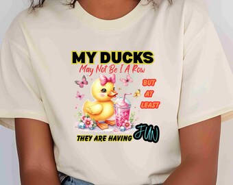 May include: Cream-colored t-shirt featuring a yellow duck with a pink bow, a pink drink, and the text "MY DUCKS May Not Be I A Row BUT AT LEAST THEY ARE HAVING FUN". The design includes butterflies and flowers.