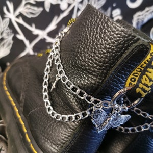 Könnte beinhalten: Schwarze Lederstiefel mit silbernen Ketten und einem Schmetterlingsanhänger. Die Stiefel haben gelbe Nähte und einen schwarzen Hintergrund.