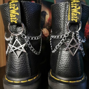 Może przedstawiać: Czarne skórzane buty z żółtymi akcentami i srebrnymi łańcuchami z gwiazdkami. Łańcuchy są przymocowane do butów srebrnymi kółkami. Buty mają grubą, czarną gumową podeszwę.