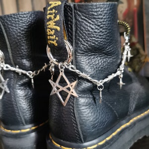 Puede incluir: Botas de cuero negro con suela cosida amarilla y una cadena plateada con abalorios en forma de estrella unidos a los cordones. Las botas tienen una correa de cuero negro con el texto "Dr. Martens" impreso en amarillo.