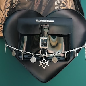 Könnte beinhalten: Schwarze herzförmige Handtasche mit einer Klappentasche und einer silbernen Schnalle. Die Tasche trägt den Markennamen "Dr. Martens" und ist mit silbernen Anhängern und einem Stacheldrahtdetail verziert. Die Tasche hängt vor einem dunkelgrünen Hintergrund.