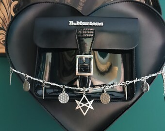 May include: Black heart-shaped handbag with a flap pocket and silver buckle. The bag features the brand name "Dr. Martens" and is adorned with silver charms and a barbed wire detail. The bag is hanging against a dark green background.
