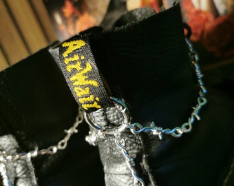 May include: Black leather boots with a yellow "AirWair" label on the back. The boots have a silver chain with barbed wire accents attached to a ring on the back of the boot.