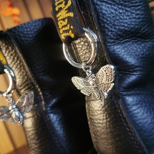 May include: Black leather boots with silver-toned moth charms hanging from the pull loops. The boots have yellow stitching and a textured surface. The moth charms are detailed with intricate patterns.