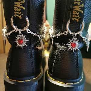 Puede incluir: Botas de cuero negro con cadenas plateadas y dijes en forma de corazón. Las botas tienen costuras amarillas y el texto "Dr. Martens" es visible en la parte posterior de las botas.